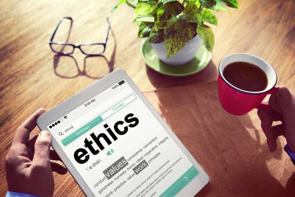 Resting on a wooden table is a tablet computer displaying an item about ethics. Beside it are spectacles, a potted plant and a coffee mug.